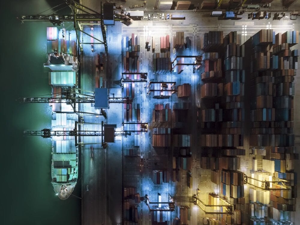 An ariel view of a port at night, featuring a cargo ship and shipping containers.