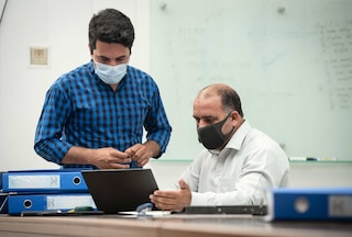 Two men wearing face masks, looking at a laptop in an office.