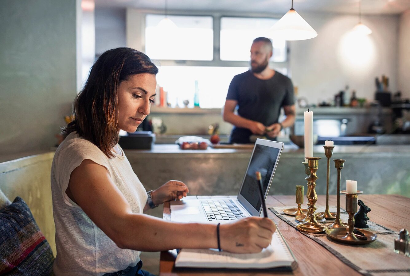 A woman sitting at a dining table with a laptop and notebook, with a man preparing food in the kitchen behind her.