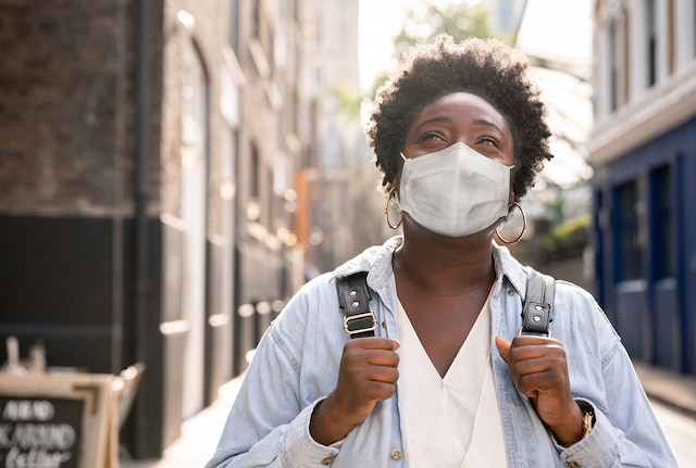 A woman wearing a face mask and holding a backpack, looking up, in a street.