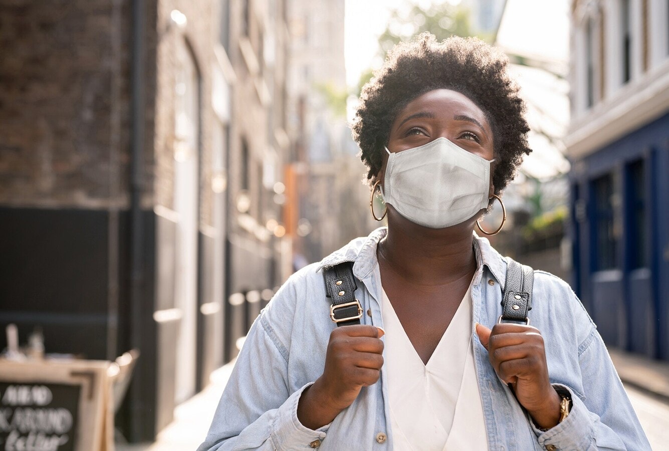 A woman wearing a face mask and holding a backpack, looking up, in a street.