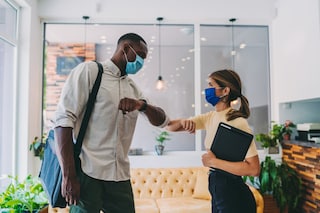 A man and a woman wearing face masks shake their elbows in an office environment.